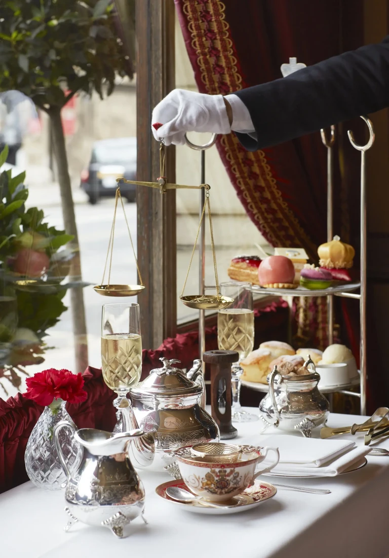 a hand in a white glove holds a golden balance above a table covered in tea dishes and a tiered pastry display