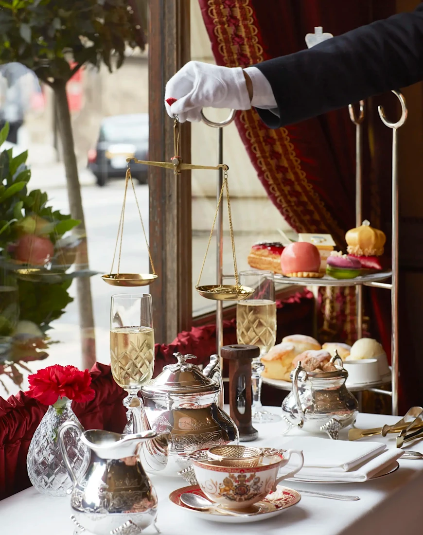 a hand in a white glove holds a golden balance above a table covered in tea dishes and a tiered pastry display