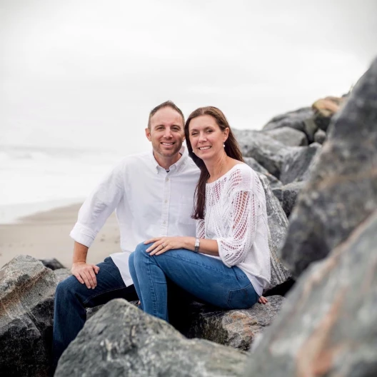 Travel Advisor Abby Rieb in a white shirt and jeans with a male, sitting on rocks by the ocean.