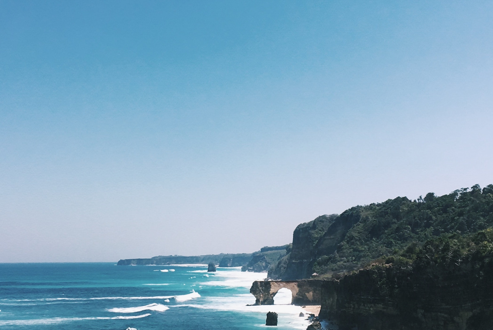 Oceanside cliff with tan dirt in Sumba, Indonesia with blue water and white surf along lush green forest.