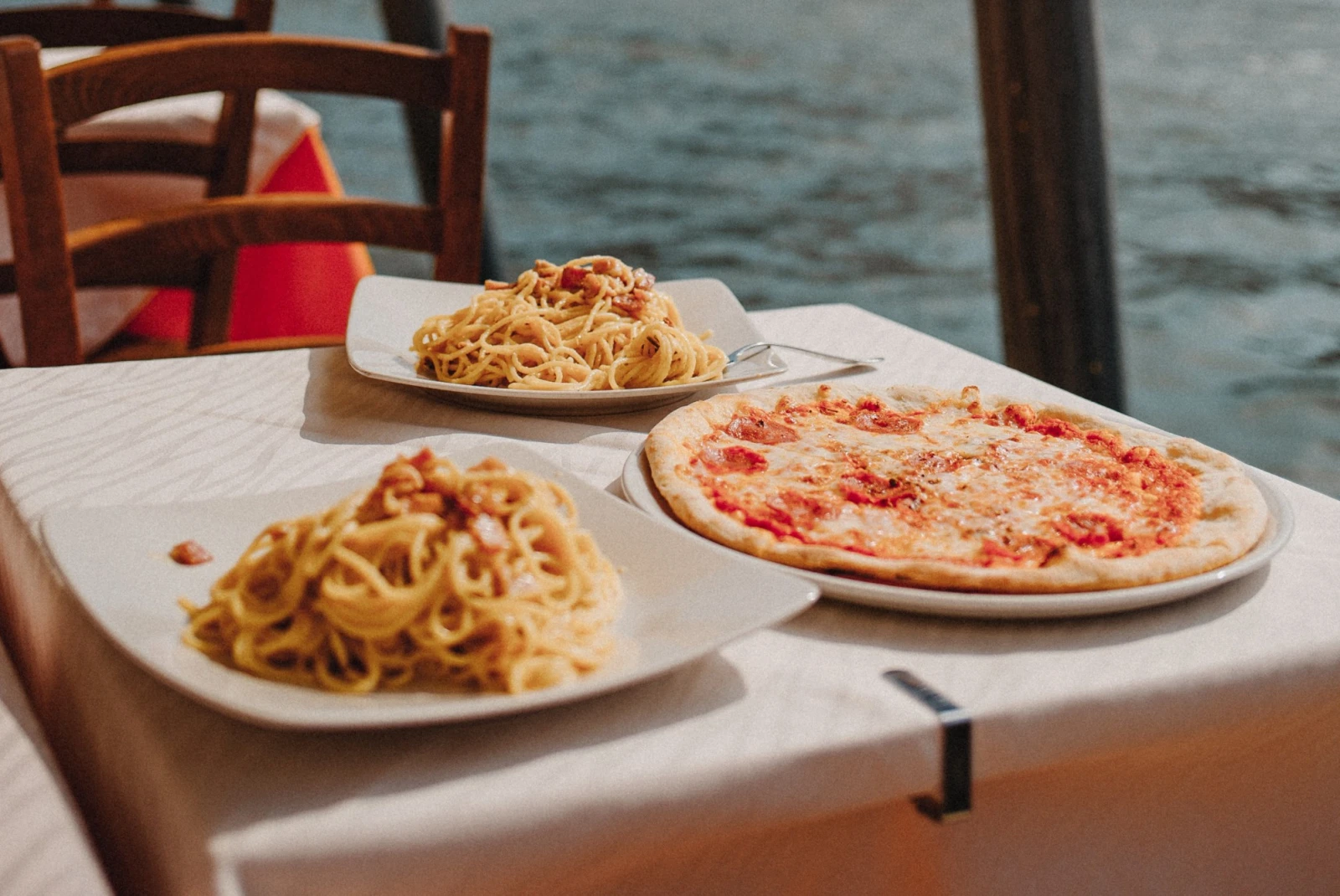 Pizza and pasta on white ceramic plates with waterbody at the background. 
