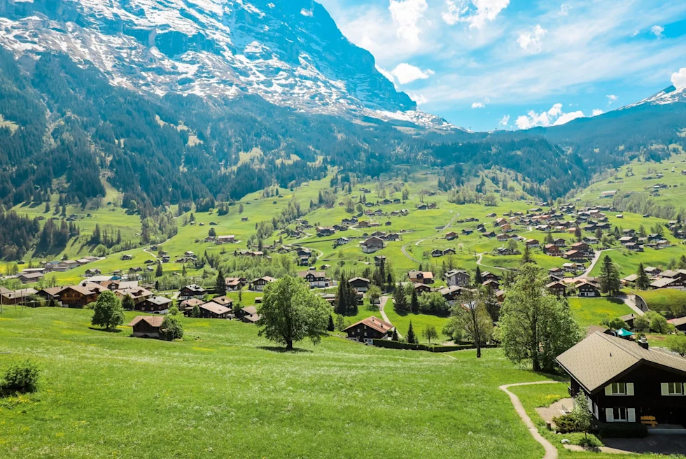 green grass valley between snow topped mountains dotted with small houses with bright blue sky
