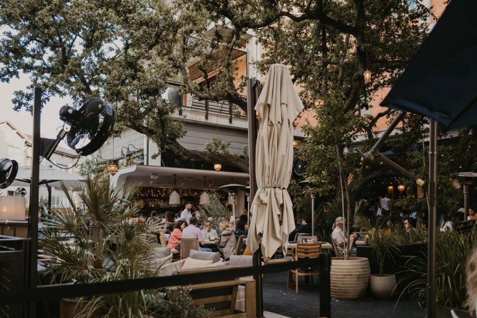 people eat and drink at an restaurant outdoor patio with trees and beige umbrellas