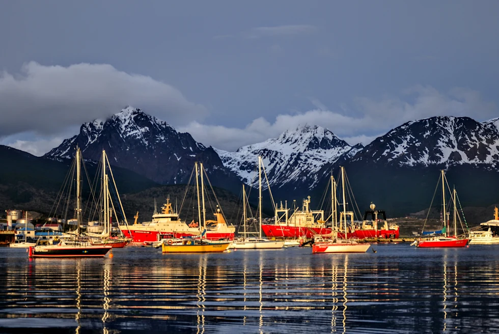 The Beagle Channel in Argentina at dusk with 15 red, orange and white boats floating in the dark waters and white snow-capped dark black mountains in the distance.