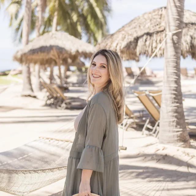 Travel advisor Hannah Breckner stands on a beach in a green dress carrying a straw hat