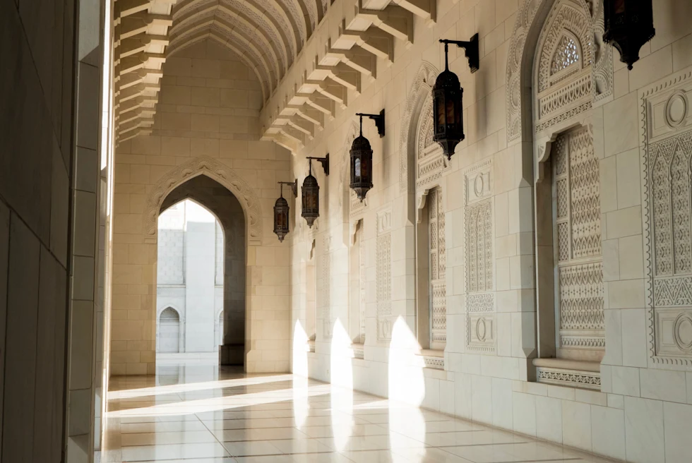 Inside of the Sultan Qaboos Grand Mosque in Oman, complete with cream stone work, beautiful arches and metal lanterns. The sun is shining in through a window against the wall on the right side.