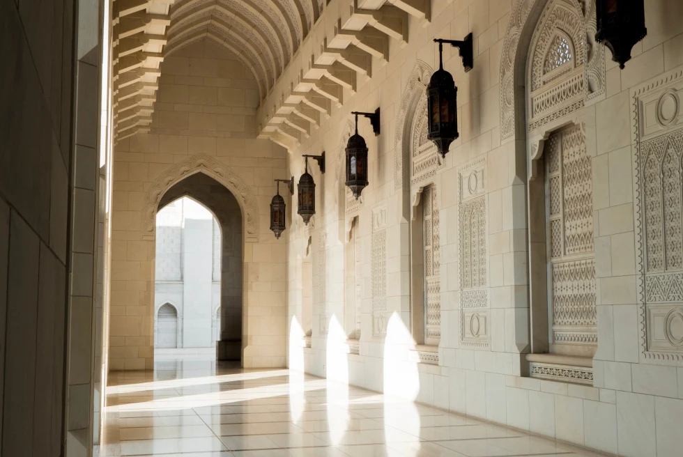Inside of the Sultan Qaboos Grand Mosque in Oman, complete with cream stone work, beautiful arches and metal lanterns. The sun is shining in through a window against the wall on the right side. 