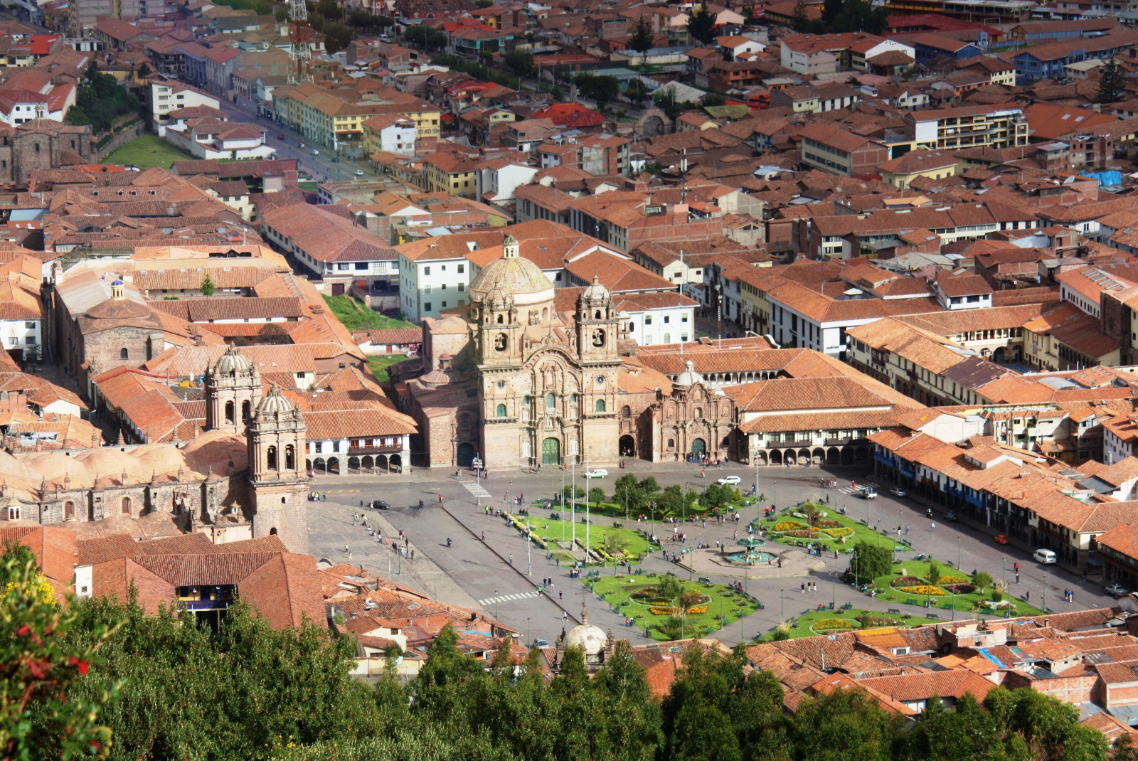 birdseye view of a cluster of tan buildings with orange roofs in cusco peru with large green grass square and trees