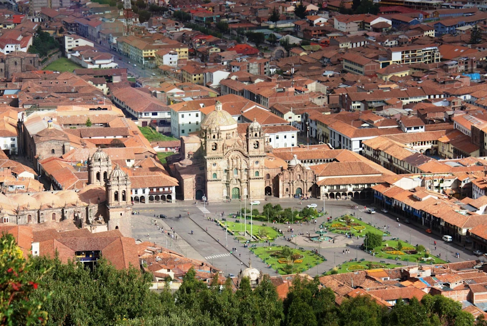 birdseye view of a cluster of tan buildings with orange roofs in cusco peru with large green grass square and trees