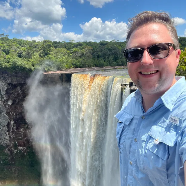 Travel Advisor Jim Platner in a blue shirt standing in front of a large waterfall and green trees.