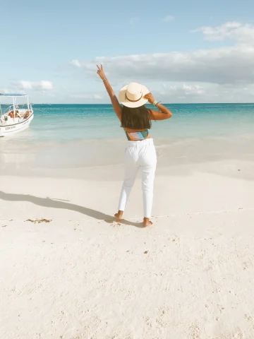 Girl with white brimmed hat stands with arm in the air by the ocean in Tulum, Mexico