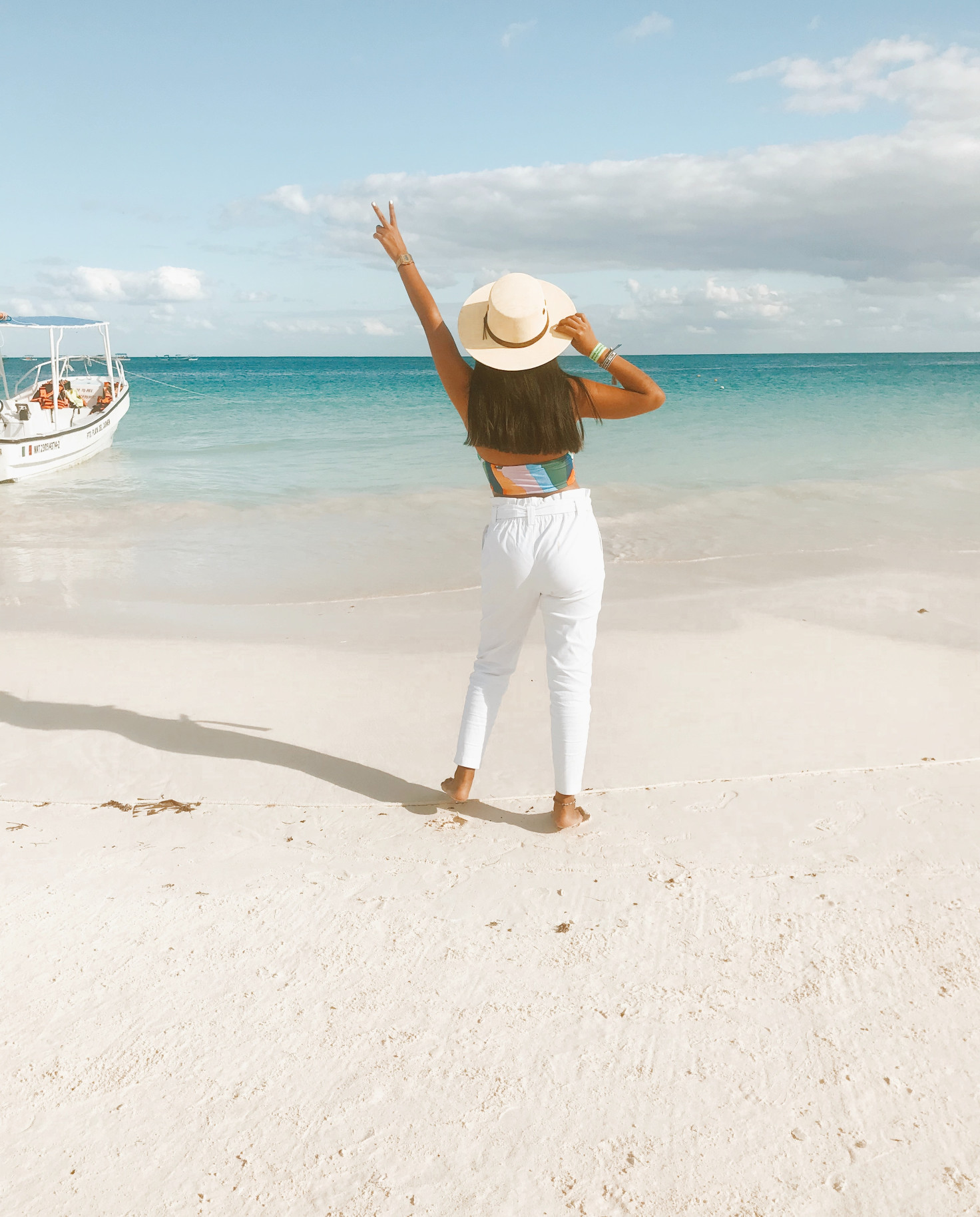 Girl with white brimmed hat stands with arm in the air by the ocean in Tulum, Mexico