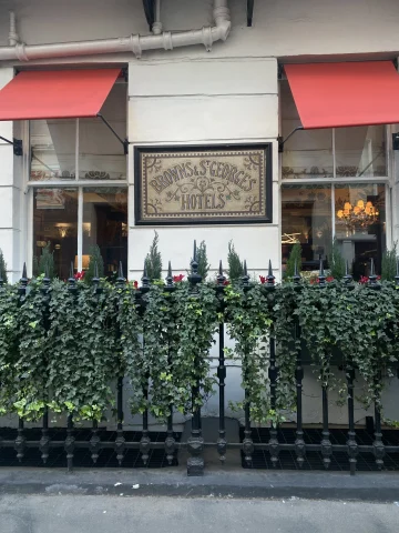 A photo of two windows with red awnings over them on either side of an old sign that reads, "Browns & St. George's Hotel" and a wrought-iron fence in front of it covered in ivy. 