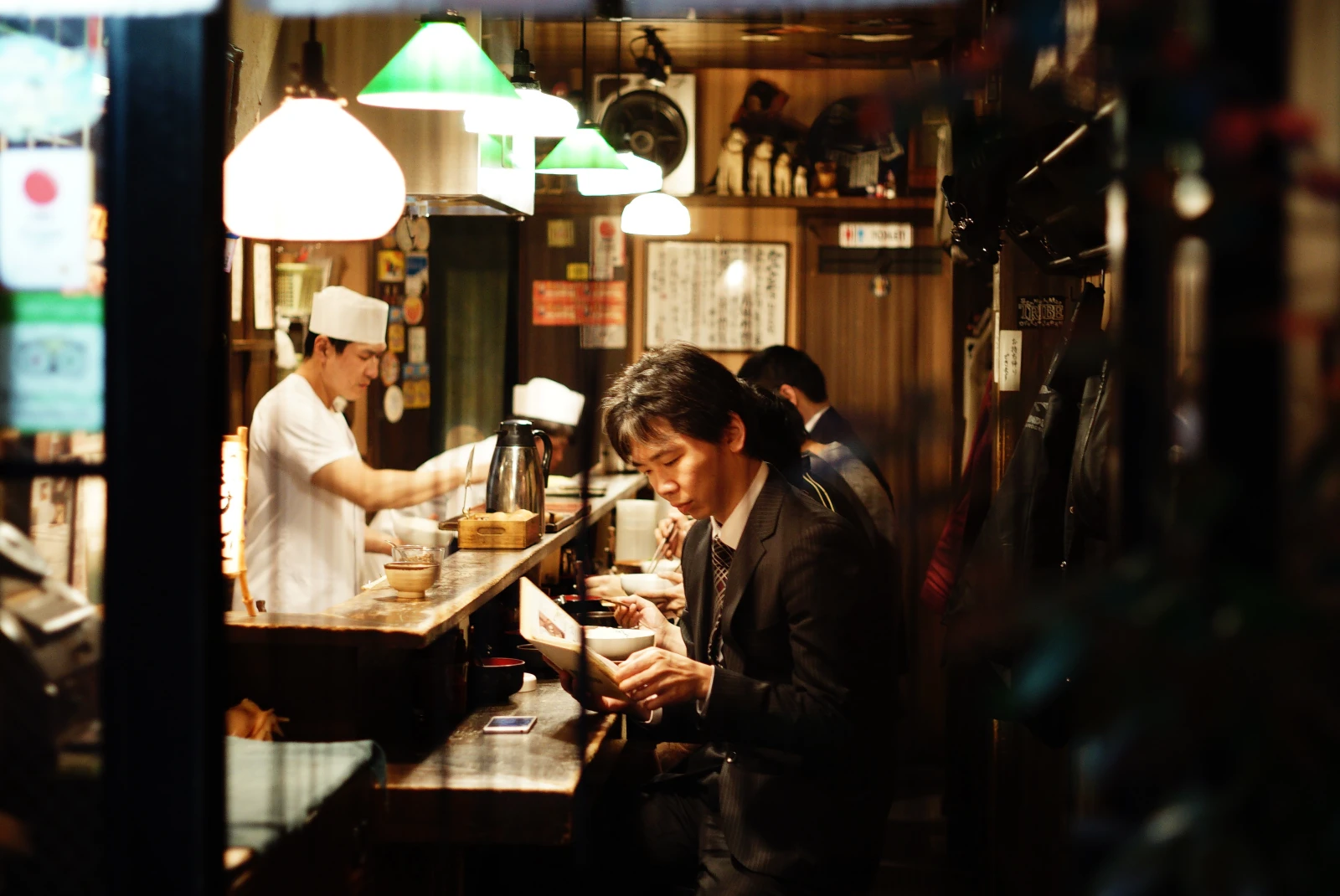 Man sitting at restaurant bar under bright lights with chef in white hat preparing food
