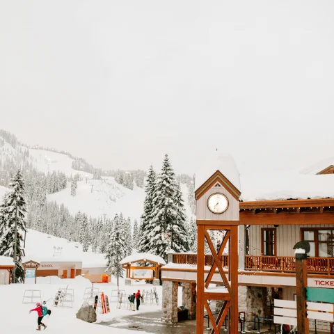 A view of a ski lodge, clock tower and people walking on the snowy terrain in the distance towards snowy pine trees and a ski hill that escapes into the fog. 