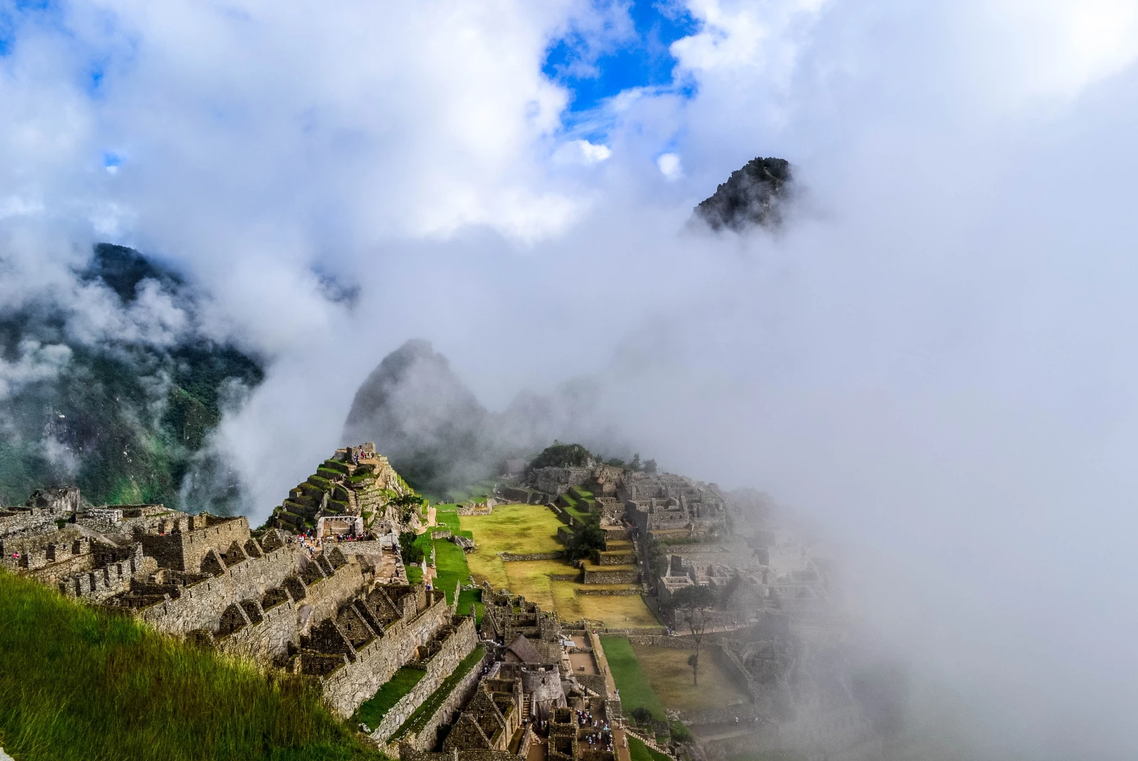 White clouds moving over Huchuy Picchu in Peru with green grass and brown stone ruins.