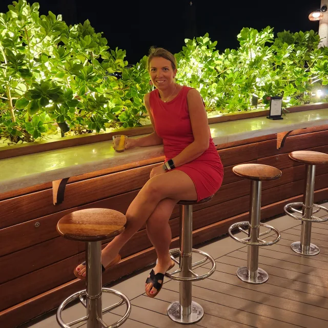 Travel Advisor Stephanie Nedeljkovic in a red dress sitting on a stool in front of green bushes.