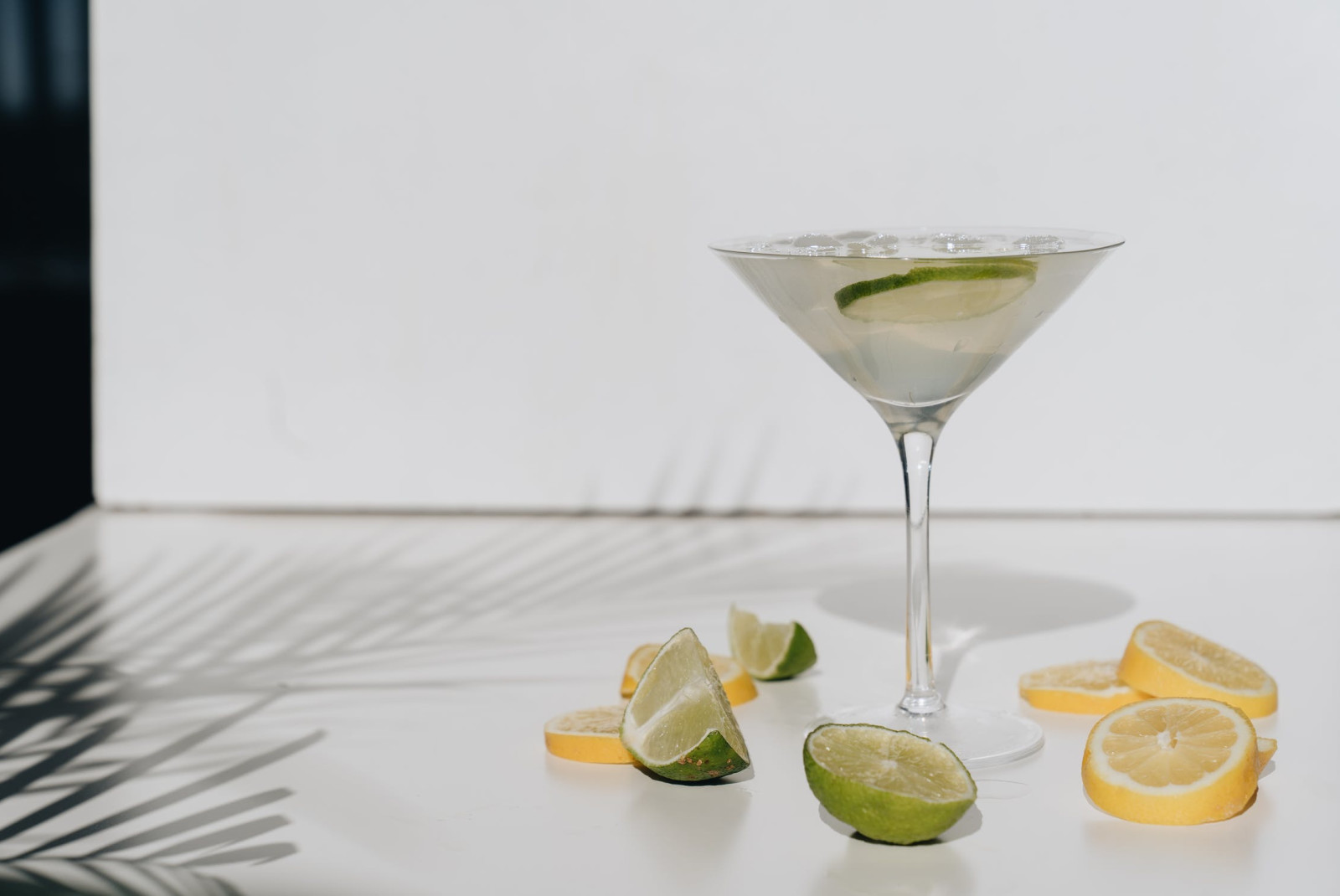 margarita in clear glass with lemons and limes on a white table with palm leaves in the shadow