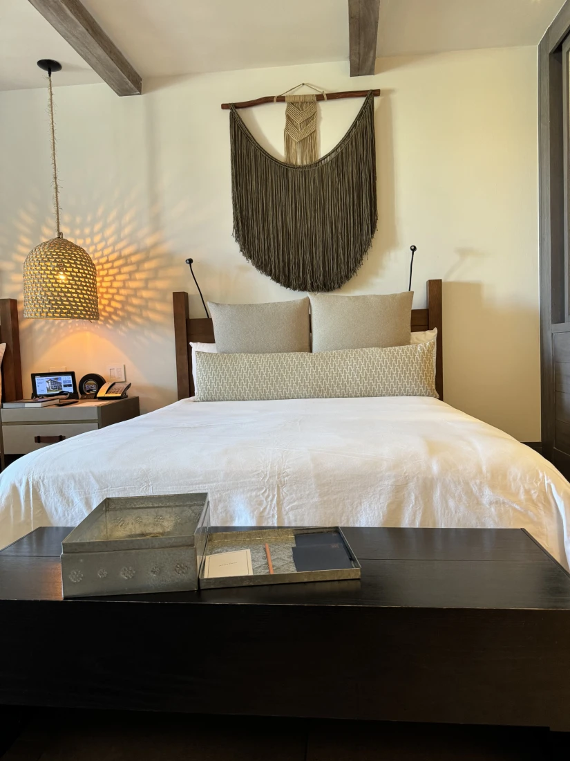 A white bed with a wooden table at the end of it with books on top of it. There is a wicker light hanging from the ceiling to the left of the bed and a decorative piece of artwork above the bed. 