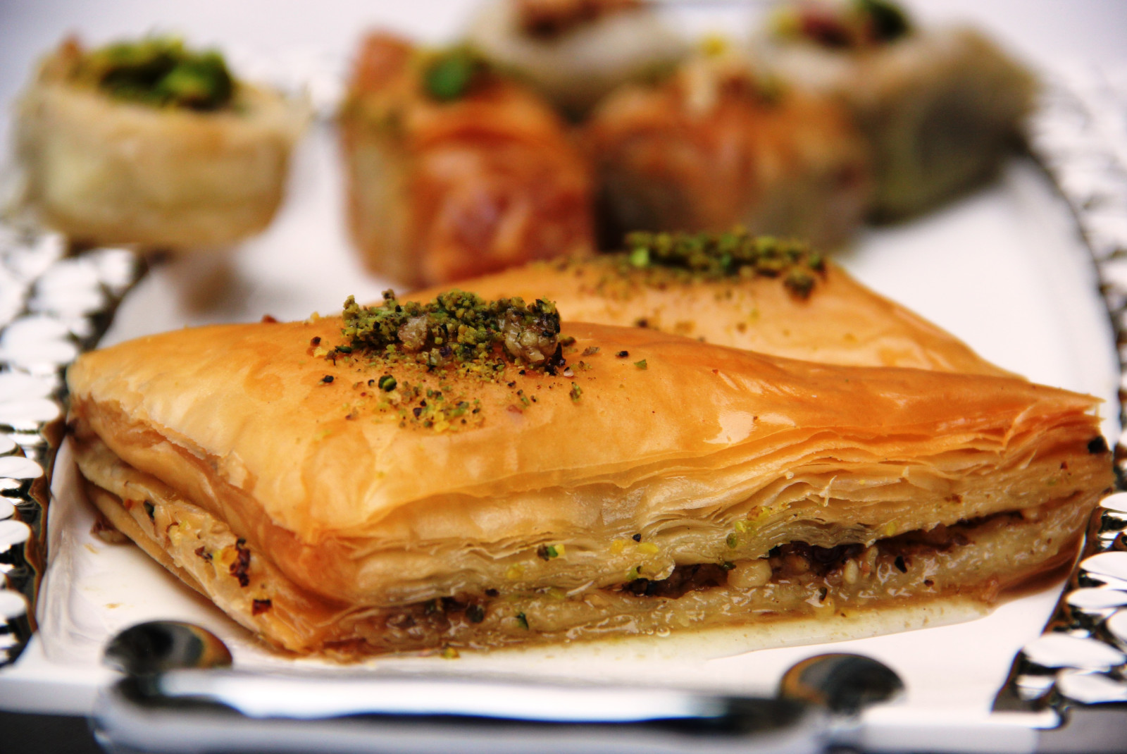 golden layered baklava pastry with pistachio crumbs on a white plate with a black lining and metal handles 