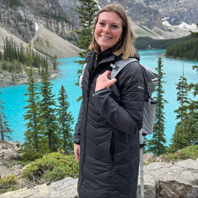 Mackenzie wearing a black jacket and backpack and posing in front of Glacier National Park 
