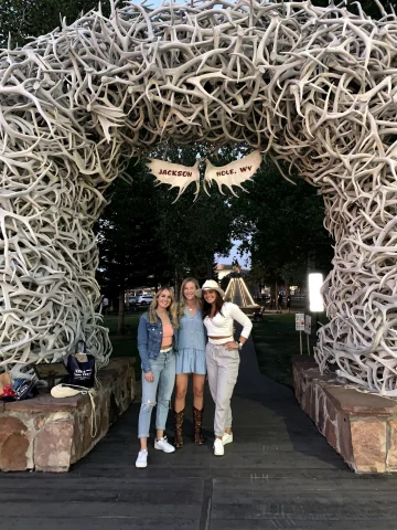 Three women standing below an outdoor archway made of antlers that says "Jackson Hole, WY" with trees in the background.