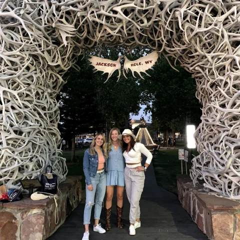 Three women standing below an outdoor archway made of antlers that says "Jackson Hole, WY" with trees in the background.