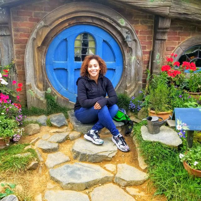 Carly Turner Travel Agent wearing blue jeans and black jacket sitting in front of brick house with blue, circular door