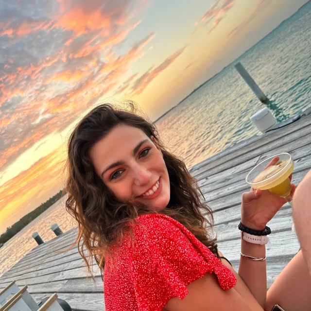 Travel Advisor Emily Buchalter with a red shirt holding a drink in front of the water at sunset.