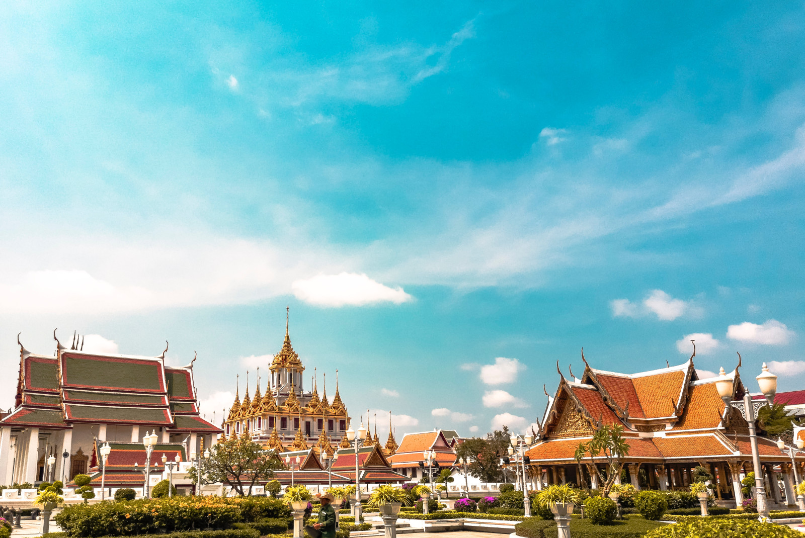 Temples in Bangkok, Thailand with a blue cloudy sky, green groomed bushes, and white paths with people walking on them. 