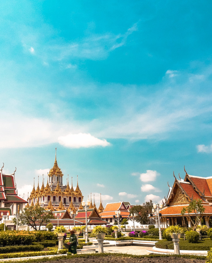 Temples in Bangkok, Thailand with a blue cloudy sky, green groomed bushes, and white paths with people walking on them. 