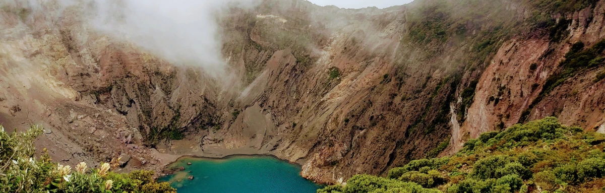 Brown crater with white wispy clouds and a blue lake with green and yellow shrubs