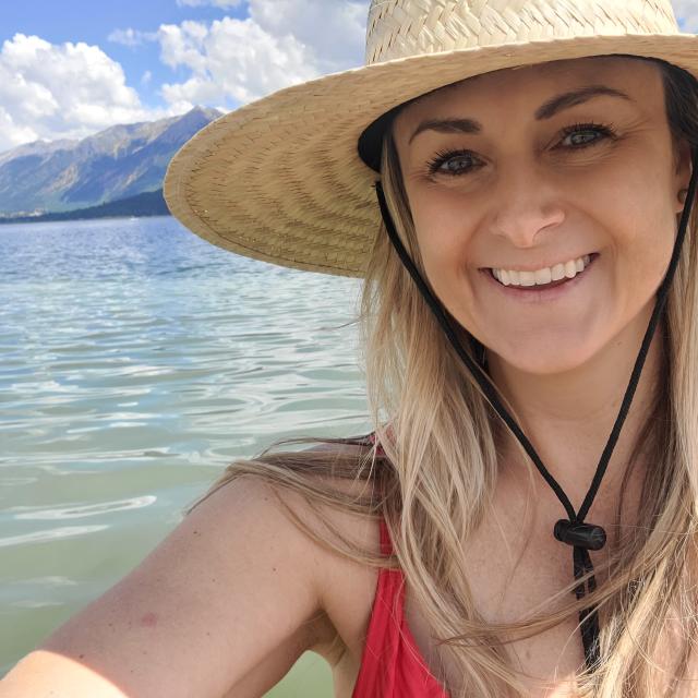 Fora travel agent Amanda Brownlie wearing straw hat and red bathing suit with water and mountains in the background