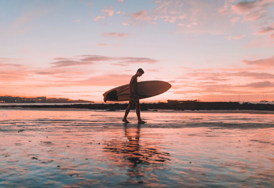 surfer walks on a beach with pink sunset