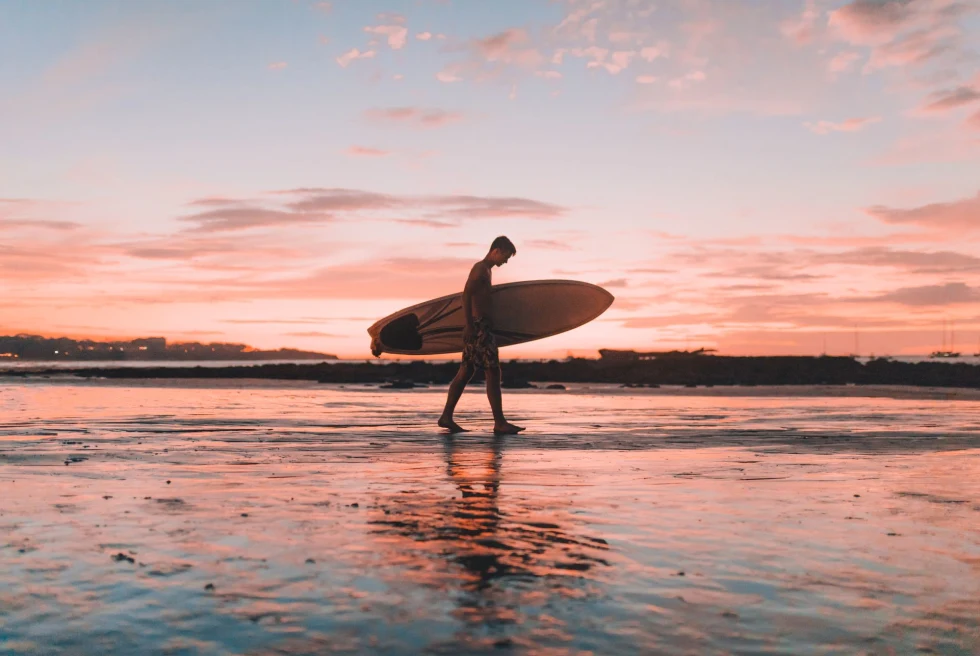 surfer walks on a beach with pink sunset