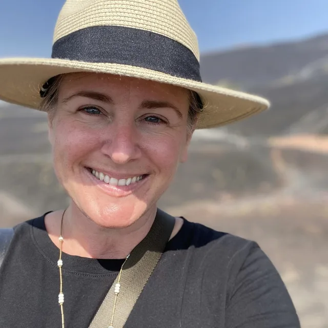 Travel Advisor Lori Loper in a black shirt and tan/black hat in front of the hills.