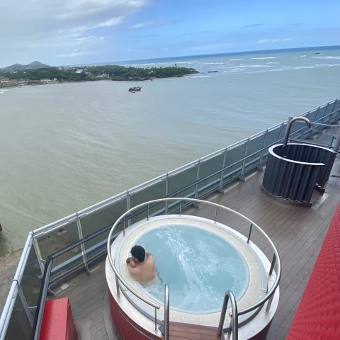 An overhead view of a person lounging in a round hot tub on a cruise ship deck, with a body of water in the background. 