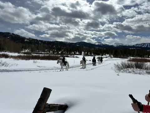 A view of four people riding horseback down a snowy trail surrounded by more snow, wild grass and mountains under a cloudy blue sky. 