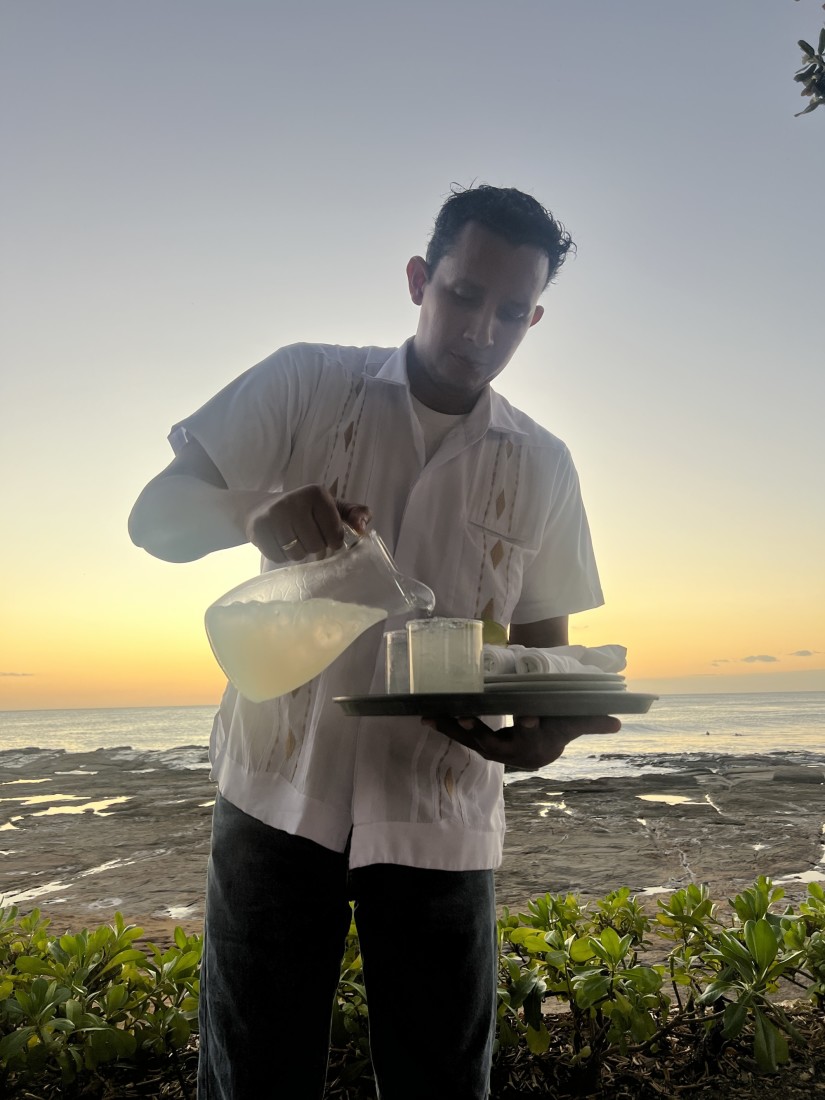Server holding a tray and pouring a drink from a jug with a sunset view of the ocean in the background