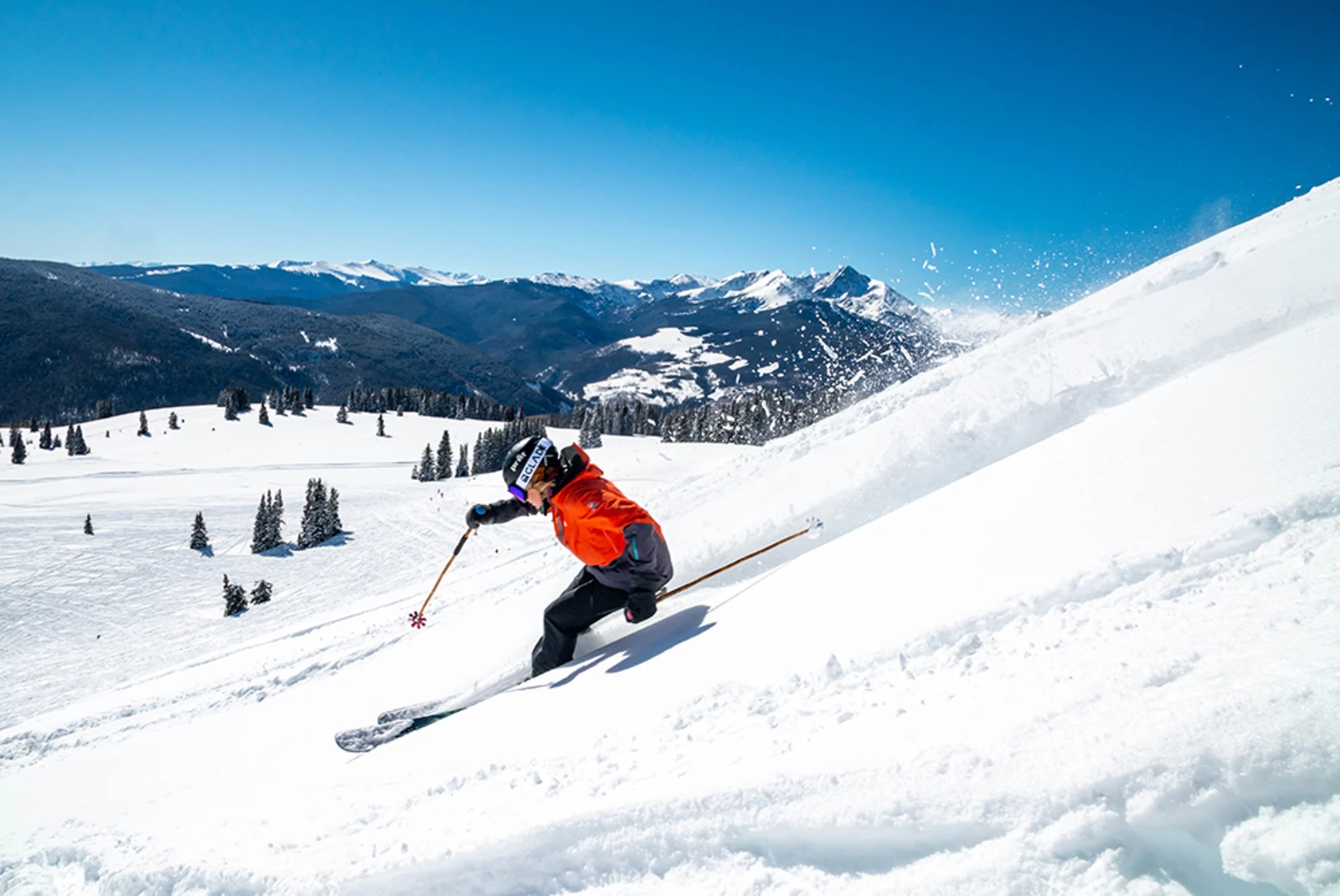 A person in red jacket, black pants and helmet skiing down a snowy mountain under a blue sky.