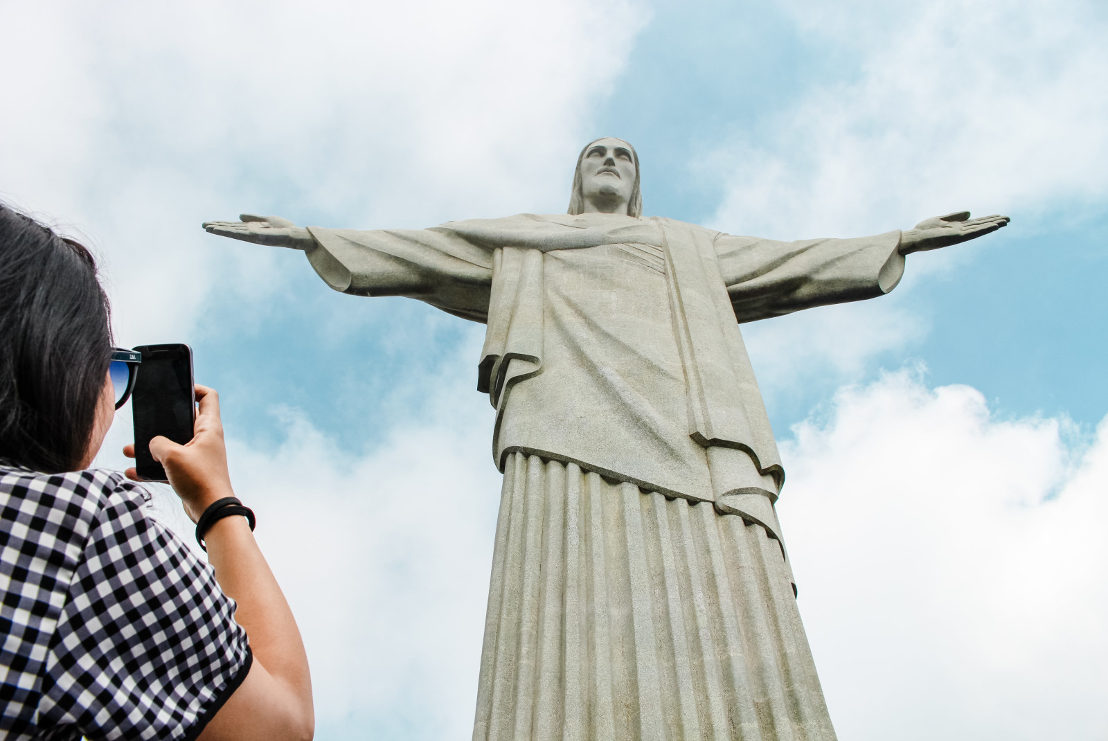 The white marbled Christ the Redeemer statue in Rio De Janeiro, Brazil with a woman in black and white checkered clothing taking a picture with an iPhone.