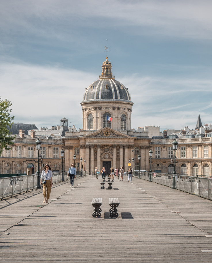 A brown bridge in Paris with people walking and a tan building with a blue rounded roof in the center with green trees