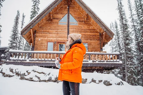 Girl in orange jacket and black pants standing in front of wooden cabin and trees in heavy snow in Stowe Vermont.