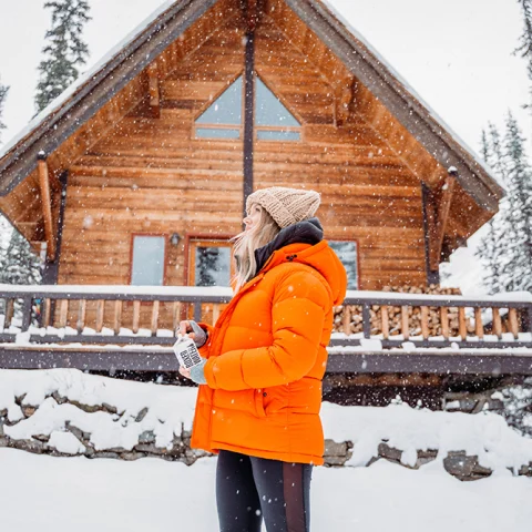 Girl in orange jacket and black pants standing in front of wooden cabin and trees in heavy snow in Stowe Vermont.