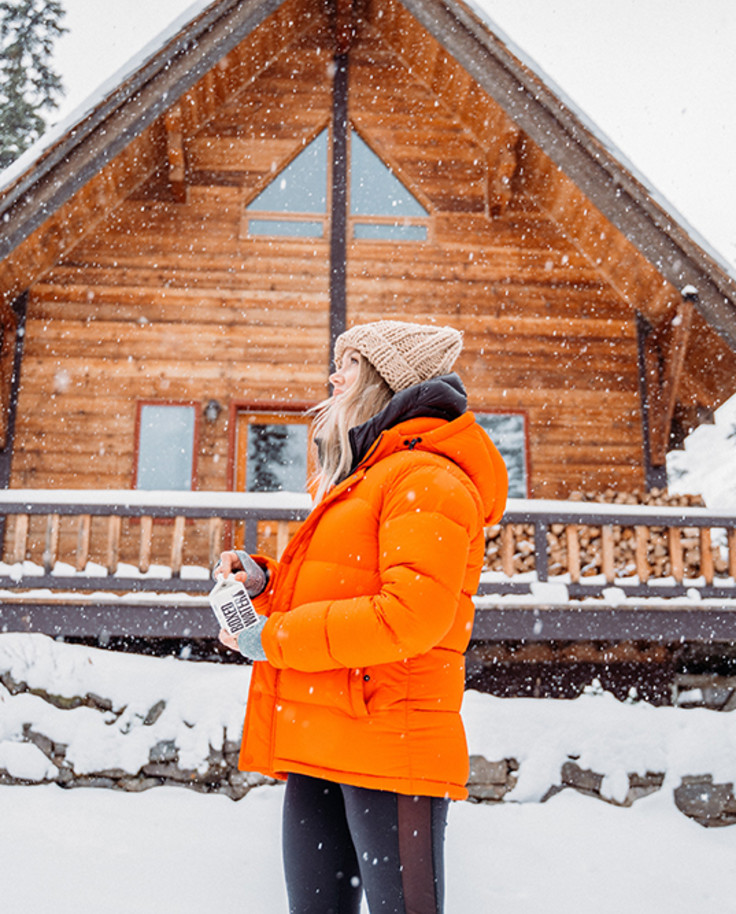 Snow covered wooden cabin and trees in Stowe Vermont with a girl in an orange jacket black pants and beige hat standing in front 