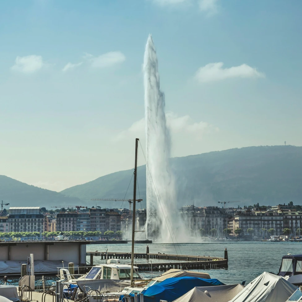 A fountain shooting water vertically up and out of the water, surrounded by buildings with mountains in the distance. 