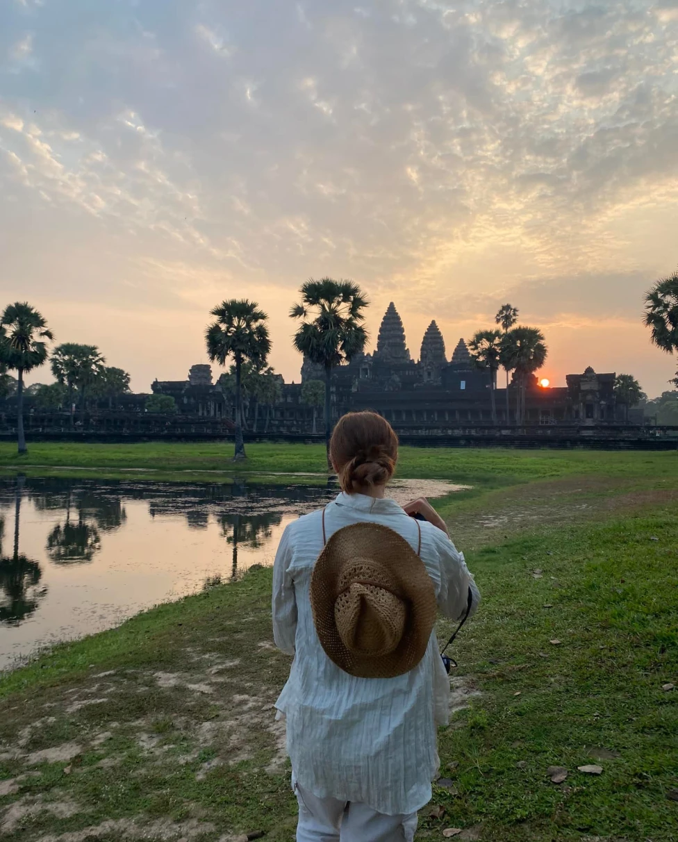  A woman with brown hat facing a building, water body and sunset.