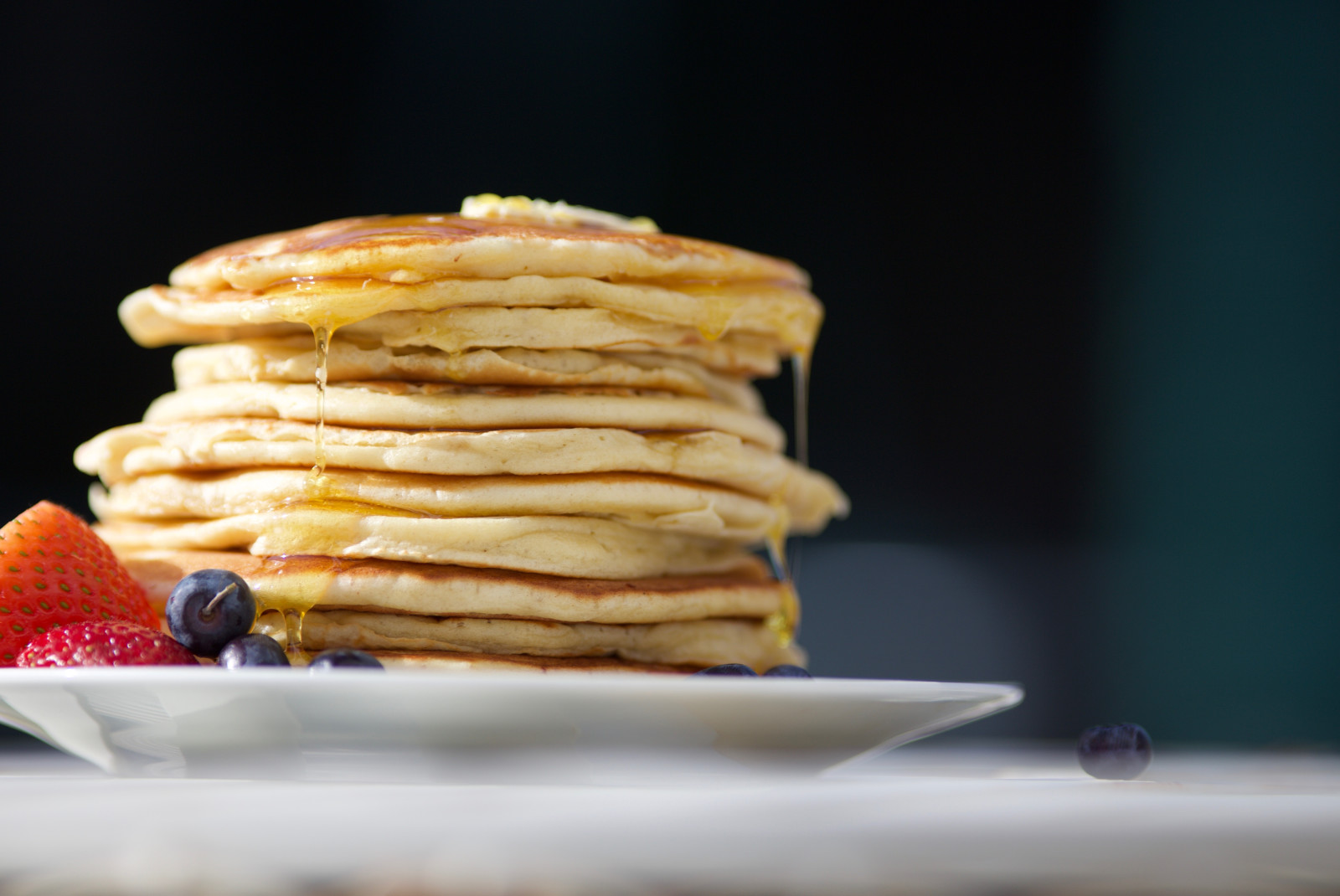 Stack of pancakes dripping with maple syrup on plate with blueberries and strawberries