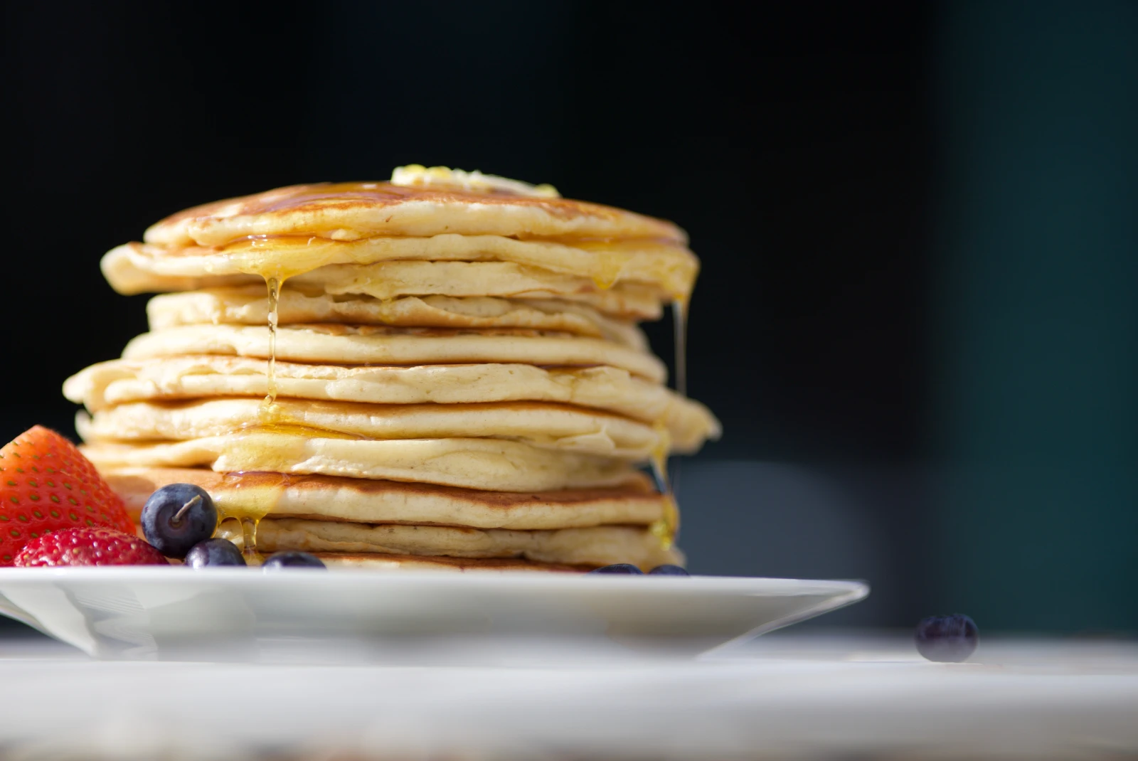 Stack of pancakes dripping with maple syrup on plate with blueberries and strawberries