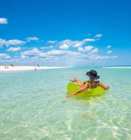 A woman relaxing on a lime green inner tube on the crystal clear water. There is a white sandy beach with people lounging in the distance. 